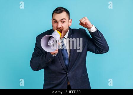 Un homme d'affaires sérieux en colère portant un costume de style officiel sombre à voix haute criant tenant le mégaphone, annonçant un message important.Studio d'intérieur isolé sur fond bleu. Banque D'Images