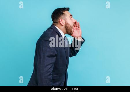 Vue latérale d'un homme barbu portant un costume de style officiel criant bruyamment la bouche largement ouverte tenant les mains sur le visage, criant annonçant son opinion.Studio d'intérieur isolé sur fond bleu. Banque D'Images