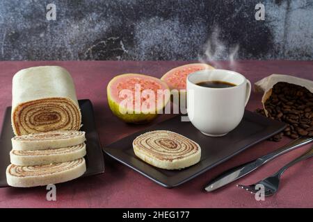 Tranche de gâteau roulé entourée de goyave, couverts, tasse et grains de café sur fond rouge. Banque D'Images