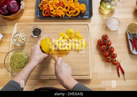 Aperçu des mains de femmes hacher du poivron jaune frais sur une planche de bois parmi les épices, les légumes et d'autres ingrédients Banque D'Images