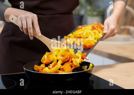 Mains de la jeune femme avec une cuillère en bois mettant le poivre jaune haché dans une poêle en position debout près d'une cuisinière électrique Banque D'Images