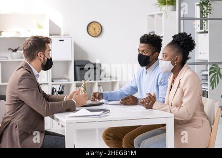 Avocat professionnel en tenue de main et masque de protection parlant à un jeune couple africain assis devant lui dans son bureau Banque D'Images