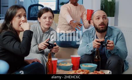 Gros plan sur des collègues jouant à des jeux vidéo sur la console, prenant des boissons et des en-cas après le travail.Collègues s'amusant avec le jeu et les contrôleurs tout en utilisant la télévision après les heures de travail. Banque D'Images