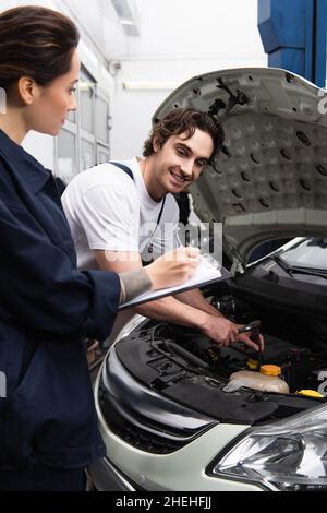 Mécanicien souriant travaillant avec la voiture près d'un collègue écrivant sur la planchette à pince en service Banque D'Images