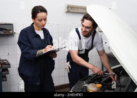 Un mécanicien écrit sur une planchette à pince près d'un collègue souriant et d'une voiture dans un garage Banque D'Images