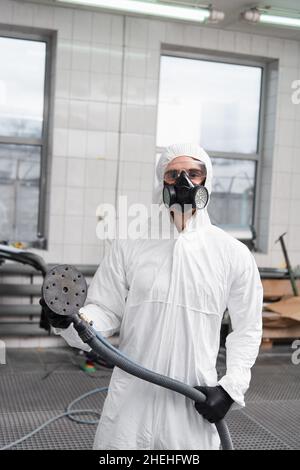 Workman en costume de noisette et respirateur tenant le polisseur de voiture dans le garage Banque D'Images