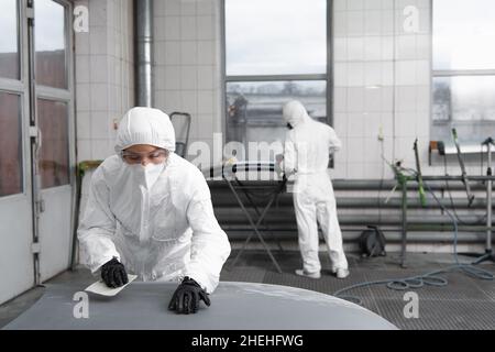 Femme de travail dans des lunettes et une combinaison de noisettes tenant le papier de verre près du capot de voiture dans le garage Banque D'Images