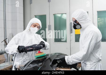 Femme de travail en costume noisette polissage de pièce de voiture près de collègue dans le respirateur en service Banque D'Images