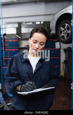 Femme de travail en gants et écriture uniforme sur le presse-papiers près de la voiture floue dans le garage Banque D'Images