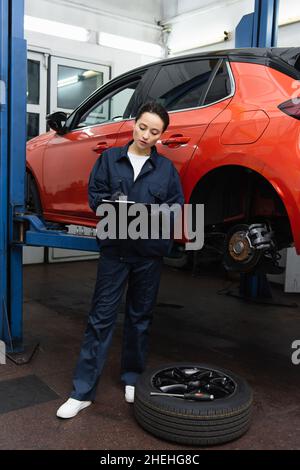 Mécanicien en écriture uniforme sur le presse-papiers près du pneu, du tournevis et de la voiture dans le garage Banque D'Images