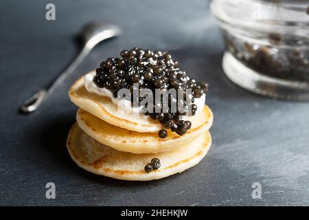 Caviar d'esturgeon noir sur de petites crêpes blini, un pot en verre et une cuillère sur fond gris foncé Banque D'Images