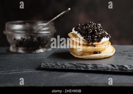 Caviar d'esturgeon noir sur de petites crêpes blinis à la crème aigre et un pot en verre avec caviar sur une ardoise Banque D'Images