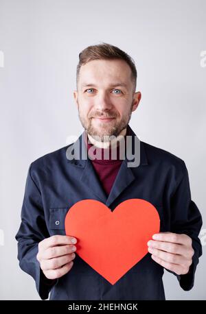 Portrait demi-longueur de jeune homme de taille basse barbu debout près du mur blanc tenant la forme de coeur rouge.Travailleur avec coeur rouge de papier.Romance, concept de relations.Saint Valentin.Image de haute qualité Banque D'Images