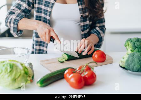 Une femme prégnante non reconnaissable a coupé le concombre sur une planche à découper en bois en mettant des légumes frais et des fruits sur une table dans la cuisine.Grossesse, maternité, santé Banque D'Images