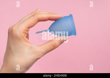 Main féminine tenant une coupe menstruelle en silicone bleu sur fond rose gros plan.Produit de soins corporels et personnels écologique et réutilisable. Banque D'Images