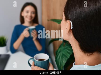 Une femme adulte atteinte d'une déficience auditive utilise une prothèse auditive pour communiquer avec sa femme lorsqu'elle boit du thé à la maison.Solutions auditives Banque D'Images