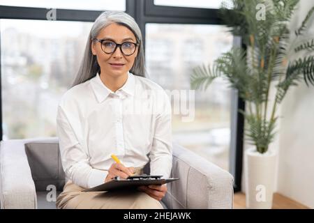 Sympathique et socialement confiante femme d'âge moyen portant des lunettes assis dans le fauteuil et regardant l'appareil photo, tient un bloc-notes et un stylo.Portrait d'affaires de femme thérapeute, consultant Banque D'Images