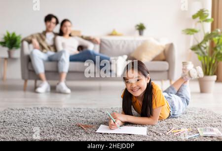 Une fille adolescente heureuse se trouve sur le sol et attire, une jeune femme asiatique et un gars se reposant sur un canapé dans le salon Banque D'Images