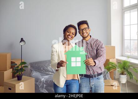 Joyeux jeune couple noir debout dans leur nouvelle maison, tenant la maison de papier et souriant Banque D'Images