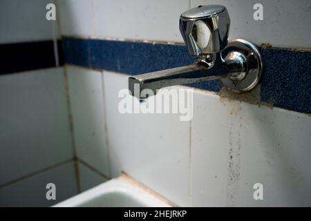 goutte à goutte, une goutte d'eau s'écoule d'un robinet métallique corrodé sale sur un mur de carreaux sale Banque D'Images