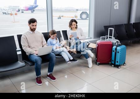 famille utilisant des appareils tout en étant assis près des bagages dans le salon de l'aéroport Banque D'Images