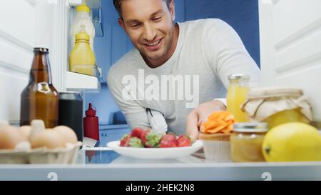 homme joyeux prenant des fraises mûres au réfrigérateur avec des boissons, des sauces, des œufs et des bocaux avec de la nourriture Banque D'Images