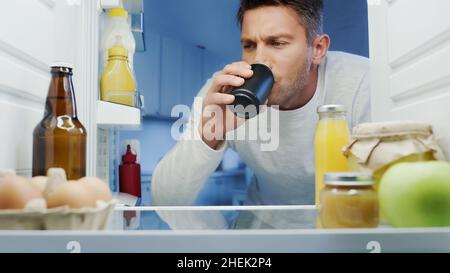homme buvant du soda en métal peut près du réfrigérateur avec des boissons, des œufs et des bouteilles avec des sauces Banque D'Images