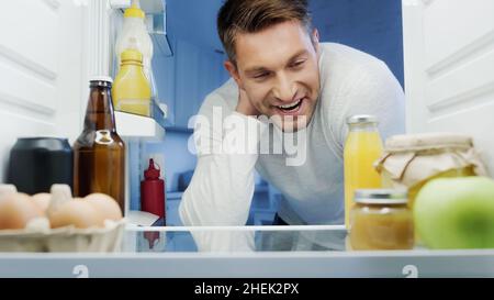 homme joyeux regardant au réfrigérateur avec du jus d'orange frais, de la bière, des œufs et des bouteilles avec sauces Banque D'Images