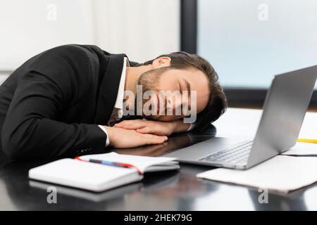 Un jeune homme d'affaires fatigué en tenue officielle fait une sieste sur le lieu de travail, un employé de bureau en costume dormant sur le bureau, prend une pause, travaille trop ou se sent ennuyer Banque D'Images