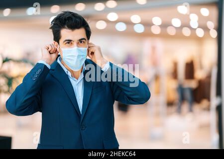 Portrait de beau jeune homme en costume élégant portant une protection masque facial regardant l'appareil photo dans le hall du centre commercial, arrière-plan flou. Banque D'Images