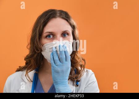 Portrait d'une femme médecin avec une droite dans les yeux, gros plan.Porter un uniforme bleu et un masque de protection sur fond orange. Banque D'Images