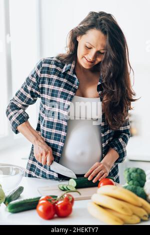 Une femme enceinte méconnaissable prépare une salade de concombre sur une planche à découper en bois avec des légumes frais et des fruits sur une table dans la cuisine.Grossesse, compagnon Banque D'Images