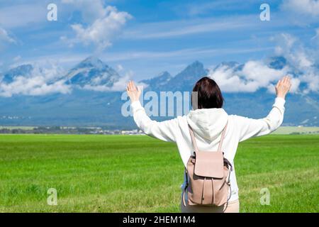 Une femme touriste dans un sweat à capuche blanc et un sac à dos bénéficie d'une belle vue sur les montagnes de Tatra tout en se tenant dans un champ vert.Les nuages flottent sur les sommets de montagne, l'espace de copie Banque D'Images