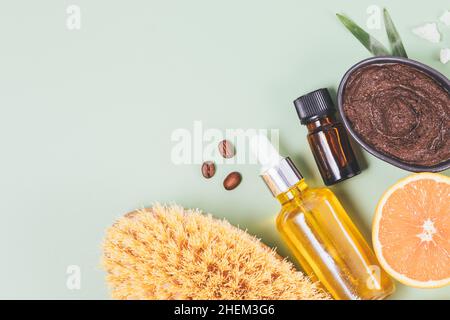 Gommage au café naturel anticellulite à côté des bouteilles d'huile essentielles, brosse de massage en bois et orange frais sur fond vert, vue du dessus Banque D'Images