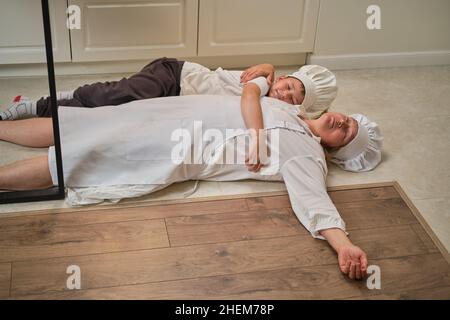 Maman et son fils drôles se détendent dans la cuisine après la cuisson. Une femme fatiguée et un garçon sont allongés sur le sol de la cuisine Banque D'Images