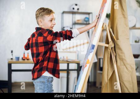 Inspiré garçon caucasien créant son chef-d'œuvre sur chevalet en utilisant des peintures et des mains.Un petit étudiant qui profite d'une leçon d'art à l'école moderne. Banque D'Images