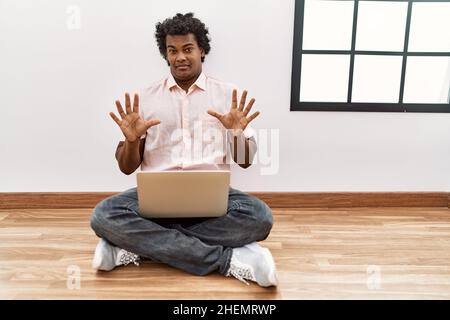 Homme africain avec des cheveux bouclés en utilisant un ordinateur portable assis sur le sol peur et terrifié avec l'expression de peur stop geste avec les mains, criant dans le choc. pa Banque D'Images