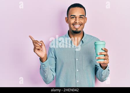 Un jeune afro-américain boit une tasse de café à emporter souriant, souriant, avec la main et le doigt sur le côté Banque D'Images