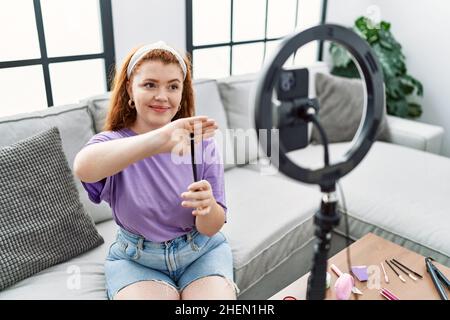 Jeune femme à tête rouge faisant le maquillage tutorial en utilisant le smartphone à la maison Banque D'Images