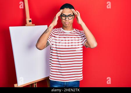 Jeune afro-américain gars debout par le peintre chevalet debout avec la main sur la tête, mal de tête parce que le stress. Souffrant de migraine. Banque D'Images