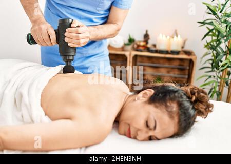 Homme et femme d'âge moyen portant un uniforme de thérapeute ayant une séance de massage du dos à l'aide d'un pistolet à percussion au centre de beauté Banque D'Images