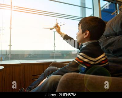 Portrait d'un adorable enfant garçon tenant un avion jouet dans une main tendue imitant le vol, contre des fenêtres panoramiques donnant sur la piste dans l'a Banque D'Images
