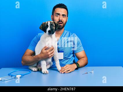 Beau vétérinaire hispanique avec la barbe de vérifier la santé de chien dans le visage de choc, regardant sceptique et sarcastique, surpris par la bouche ouverte Banque D'Images