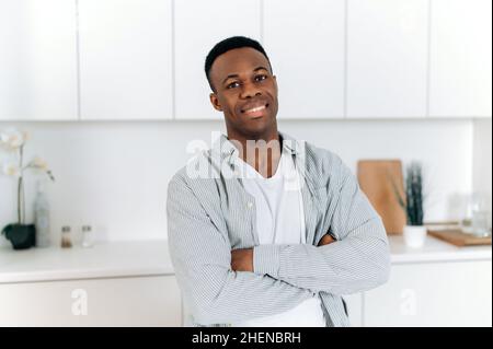 Portrait d'un homme beau afro-américain dans l'appartement.Noir homme attrayant, habillé dans des vêtements décontractés élégants, se tient à la maison dans la cuisine avec des bras croisés, regarde l'appareil photo, sourire amical Banque D'Images