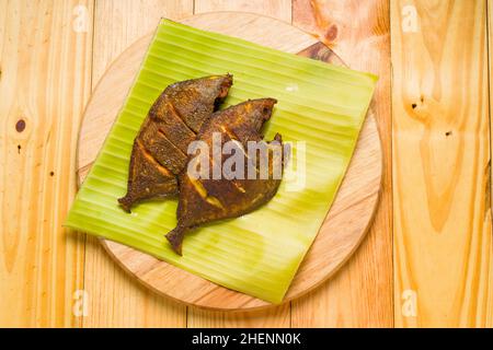 Poêlées de tranches de tomate, d'oignon et de citron, les frites de poisson Pomfret sont disposées dans une feuille de banane placée sur une base en bois. Banque D'Images