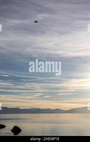Une silhouette d'avion minuscule et noire dans un grand ciel sur la surface lisse du lac Tahoe par un jour brumeux Banque D'Images