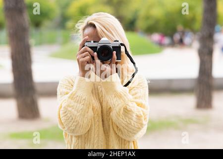 Femme blonde avec un chandail jaune prenant une photo avec un appareil photo numérique dans un parc Banque D'Images