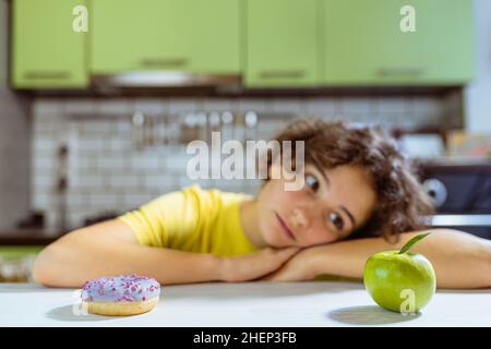 Enfant adolescent triste confronté à un choix entre le beignet et la pomme verte. Jeune fille adolescente avec un visage triste, des contusions de malnutrition sous ses yeux, est à l'alimentation Banque D'Images