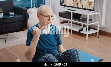 Gros plan des poids de levage des retraités et assis sur un tapis de yoga à la maison.Homme à la retraite faisant de l'exercice physique avec des haltères pour entraîner les muscles des bras et faire de l'activité d'entraînement.Personne âgée Banque D'Images