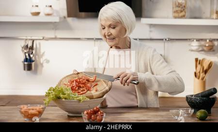 femme âgée souriante ajoutant du poivron en tranches à la laitue tout en préparant une salade de légumes Banque D'Images